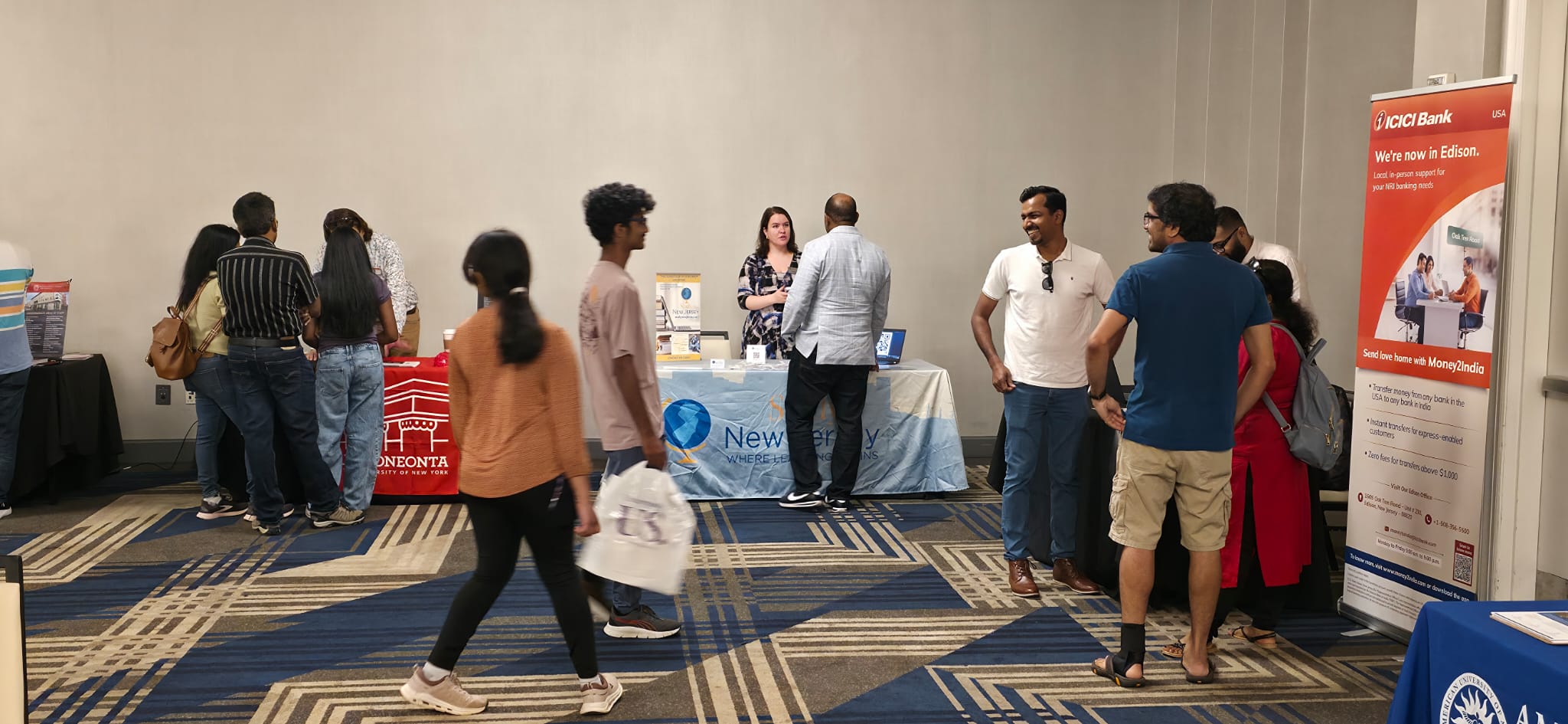 Attendees walking past the ICICI Bank, Study NJ, and SUNY Oneonta tables, with an American University of Antigua representative discussing cross-collaboration programs